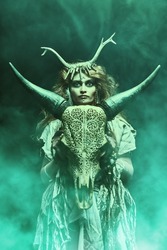 Female shaman in an ethnic dress doing a mysterious ritual with a help of an animal skull. Dark mystical background with green haze. Black magic concept, fantasy. Paganism. Halloween.
