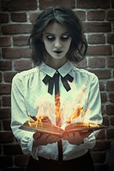 Portrait of a poltergeist girl with colorless eyes standing in a gloomy room by a brick wall with an opened burning book. Halloween. Horror scene. 