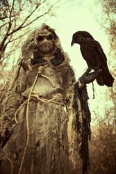 An evil Scarecrow with a canvas bag on his head and in a sackcloth robe stands in a dense forest with a black raven on his hand. Terrible forest sorcerer. Halloween Tales. Horror, thriller.