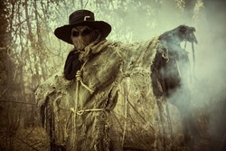 Halloween. A scene of horror, when an evil terrible Scarecrow with a bag on his head, in a sackcloth vestment and a black hat, stands in the thicket of the forest, surrounded by fog. Horror, thriller.