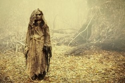 A creepy Scarecrow with a canvas bag on his head, in a burlap robe stands in a dense forest. Vintage style. Halloween Tales. Horror, thriller.
