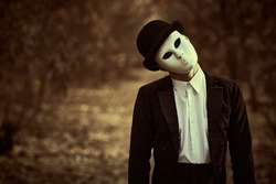 Portrait of a mysterious gentleman in a white carnival mask, bowler hat and black tailcoat standing in an old autumn park. Mime, carnival. Halloween. Vintage style. Copy space.