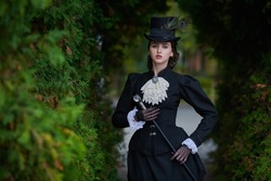 Historical reconstruction of the late 19th and early 20th centuries. Elegant brunette lady in a strict black dress posing in an autumn park. Historical makeup and hairstyle.