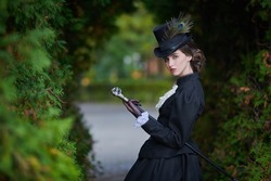A confident and sophisticated lady in a strict black suit of the 19th century poses with a walking stick against the backdrop of an autumn park.