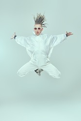 Full length shot of a modern punk rock musician with dreadlocks in white overalls and stylish glasses  jumping on a light background with his arms streche aside. Youth alternative culture. 