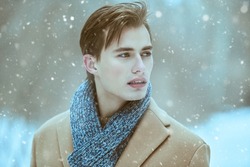 Fashion shot of a stylish handsome young man in a trendy coat and scarf posing outdoor on a snowy day. Men's beauty and fashion.