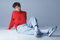 Handsome guy model in a red sweater, light jeans and and  sneakers posing sitting on the floor in the studio. Men's youth fashion. 