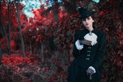 Historical reconstruction of the late 19th and early 20th centuries. Elegant brunette lady in a strict black dress posing in the background of red foliage. Historical makeup and hairstyle.