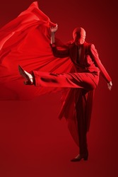 Fashionable tones. Studio portrait of a female fashion model posing in motion in a red pantsuit and red cloth covering her face, holding red fabric fluttering on a red background.