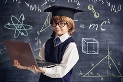 Portrait of a clever little boy in school uniform and glasses holding a laptop in the background of a blackboard with scientific formulas and diagrams. Smart children. Education. 