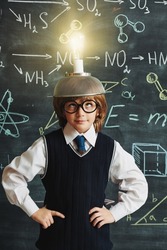 A cute smart boy in neat school uniform and glasses stands at the blackboard with chemical formulas with a strange invention on his head. Clever kids. Eureka. Educational concept. 