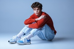Handsome guy model in a red sweater, light jeans and sneakers poses sitting on the floor in the studio. Men's youth fashion. 