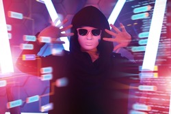 A portrait of teenage boy in a black hoodie, mask and black sunglasses hiding his face works with holographic data on virtual screen. Studio shot among neon lights. Teen hacker in cyberspace. 
