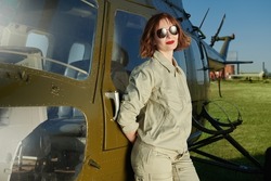 Portrait of an attractive professional woman pilot with bright red lips and black sunglasses standing by a helicopter.