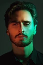 A portrait of a masculine brunet man in a black shirt looking calmly and thoughtfully at the camera. Dark background lit by multicoloured lighting. Male beauty.