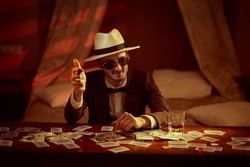A joyful gambling man in elegant suit, hat and sunglasses sits in a bedroom at a table playing cards for money and smoking a cigar. Retro style. Mafia, gambling and the criminal world.