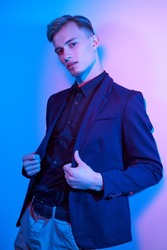 A well-groomed handsome young man dressed in a smart black shirt, jacket and trousers standing by the wall in multicoloured lighting. Male fashion.
