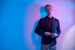 Male fashion. A handsome young blond man wearing a smart shirt, jacket and trousers stands by a white wall in multicoloured lighting and joyfully smiles. Copy space.