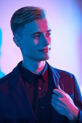 Male fashion. A handsome young blond man wearing a smart shirt, jacket and trousers stands by a white wall in multicoloured lighting and joyfully smiles. 