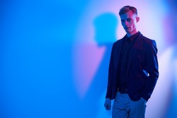 A handsome well-groomed blond man wearing a smart shirt, jacket and trousers stands by the wall in multicoloured lighting and smiles. Male fashion. Copy space.