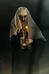 Scary devilish possessed nun standing with a candle in a dark room. Horrors and Halloween.