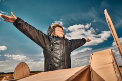 A happy little boy in a leather jacket and aviator's helmet imagines that he is flying in a cardboard plane through a blue sky. Children's games and dreams. 