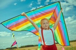 Carefree summer holidays. A cute red-haired girl in summer clothes flies a multicoloured kite in an open field. Summertime. Children's games. Happy childhood.