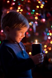 Little toddler boy holding candle with colorful lights from Christmas tree on background.