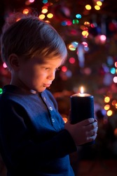 Little toddler boy holding candle with colorful lights from Christmas tree on background.
