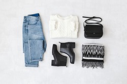 Blue jeans, white knitted sweater, small black cross body bag, leather ankle boots and striped scarf on grey background. Overhead view of woman's casual day outfits. Trendy hipster look. Flat lay.