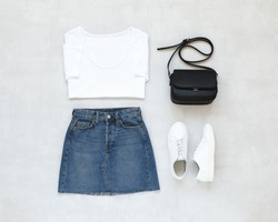 White t-shirt, blue denim mini skirt, small black cross body bag, white sneakers on grey background. Overhead view of woman's casual outfit. Flat lay, top view. Trendy simple basic minimalistic look.