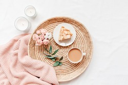Wicker tray, cup of coffee with milk, piece of cake, rose flowers, eucalyptus branch, candles, pink knitted plaid or blanket. Breakfast in bed. Stylish home interior decor. Flat lay, top view.