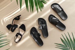 Summer women's shoes. Black heeled sandals, flat sandals, rubber slippers and tropical palm leaves on beige background. Trendy female footwear. Flat lay, top view.