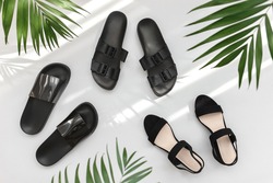 Summer women's shoes. Black heeled sandals, flat sandals, rubber slippers and tropical palm leaves on grey background. Trendy female footwear. Flat lay, top view.