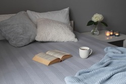 Stylish cosy bedroom in grey colors. Cozy interior, home decor. Bed with grey blanket, pillows, knitted plaid, bedside table, vase with hydrangea flower, candles, book and a cup of tea.