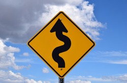 Winding road, yellow and black warning sign with curvey arrow against a cloudy sky 