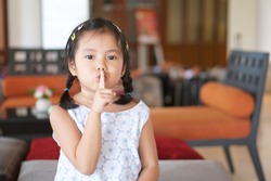Asian child cute or kid girl squat and forefinger close mouth for tell quietly shh or stop silence and secret surprise with play hide and seek in library room at school or reception room