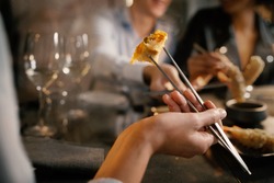 Detail of a woman's hand grabbing a spring roll with metallic chopsticks. He sits at a table with friends in a fancy asiatic fusion food restaurant.