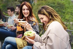 Gathering of best friends eating salads in plastic-free bowls, talking, joking and having fun together sitting outdoors in the park