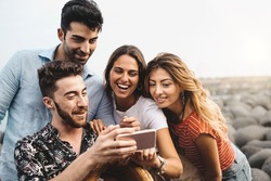 Group of cheerful young people watching social network funny content on a smartphone screen - caucasian friends get together outdoors using modern cellphone and having fun together
