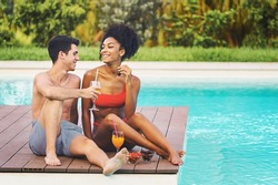 Multiethnic couple with two young romantic people relaxing sitting by the luxury swimming pool eating fruits and drinking juicy orange smoothies - Biracial couple flirting sitting poolside