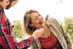 A young curvy woman laughs as she eats skewers of meat from her friend's hands during a trip to the countryside. Young adults people having fun together at picnic - people lifestyle concept
