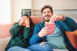 Scared millennial newlywed couple watching horror movie on tv, frightened young woman covering eyes and man laughing and eating popcorn during a thrilling scary film moment sitting on sofa at home