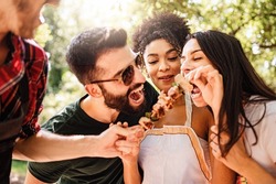 Happy multiethnic millennials playing together eating skewers and eating together skewers in the countryside at picnic - focus on African American woman - people, food and drink lifestyle concept