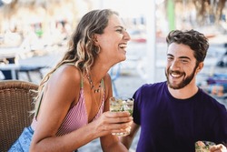 Joyful Woman and Bearded Man at Beach Bar: Close-up of a light brunette woman holding a mojito, sitting with a bearded man, both laughing heartily together.