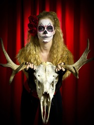 Portrait shot of a scary woman wearing sugar skull holding animal skull over red background.