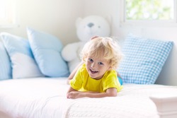 Child playing in bed in white sunny bedroom with window. Kids room and interior design. Baby boy at home. Bedding and textile for children nursery. Kid with toy and book. Nap and sleep time.