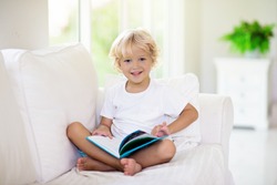 Child reading book. Kids read books. Little boy sitting on white couch in sunny living room watching pictures in story book. Kid doing homework for elementary school or kindergarten. Children study.