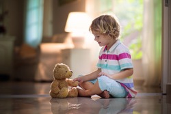 Child playing with teddy bear. Little boy hugging his favorite toy. Kid and stuffed animal at home. Toddler sitting on the floor of living room with big window at sun set. Kids play indoors.