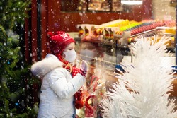 Child in face mask at store window on traditional Christmas market in coronavirus outbreak. Covid-19 pandemic. Safe Xmas shopping, gifts and presents with kids on snowy winter day. 